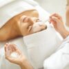 Woman enjoying facial treatment at beauty parlor or spa salon. Professional beautician holding small bowl with white clay mask and using brush to apply it on face of young woman client