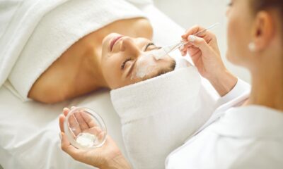 Woman enjoying facial treatment at beauty parlor or spa salon. Professional beautician holding small bowl with white clay mask and using brush to apply it on face of young woman client