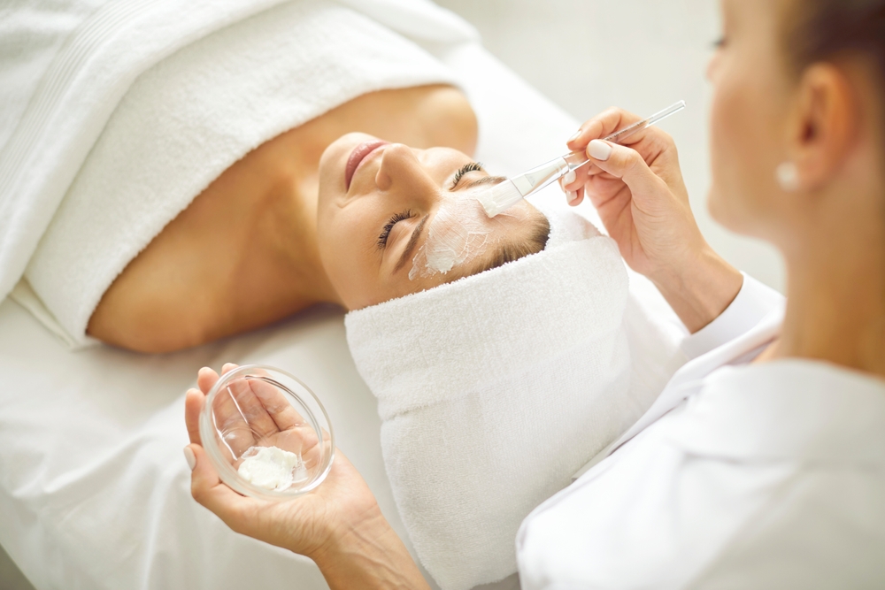 Woman enjoying facial treatment at beauty parlor or spa salon. Professional beautician holding small bowl with white clay mask and using brush to apply it on face of young woman client